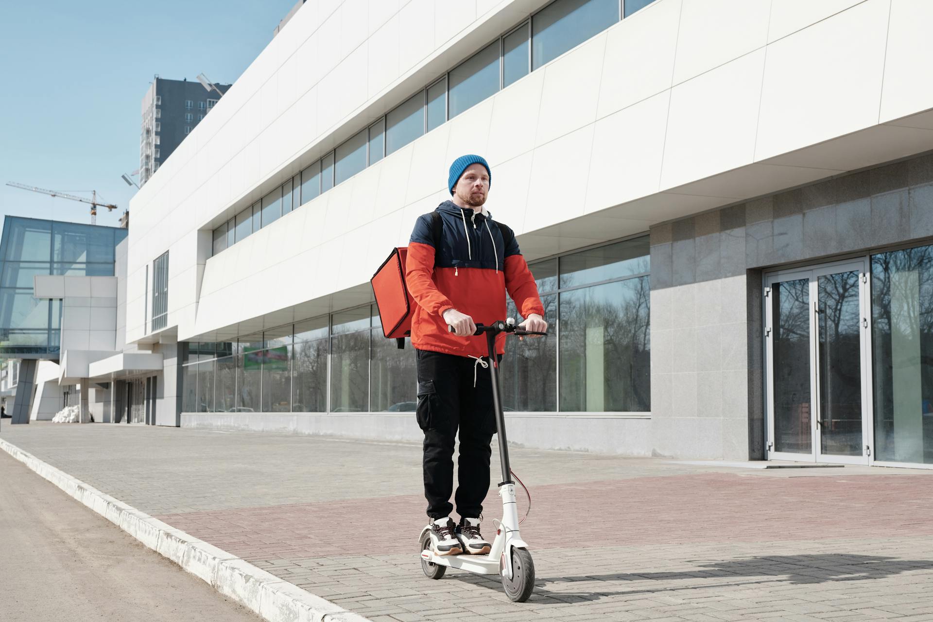 A male courier on an electric scooter delivering food outside a modern building.