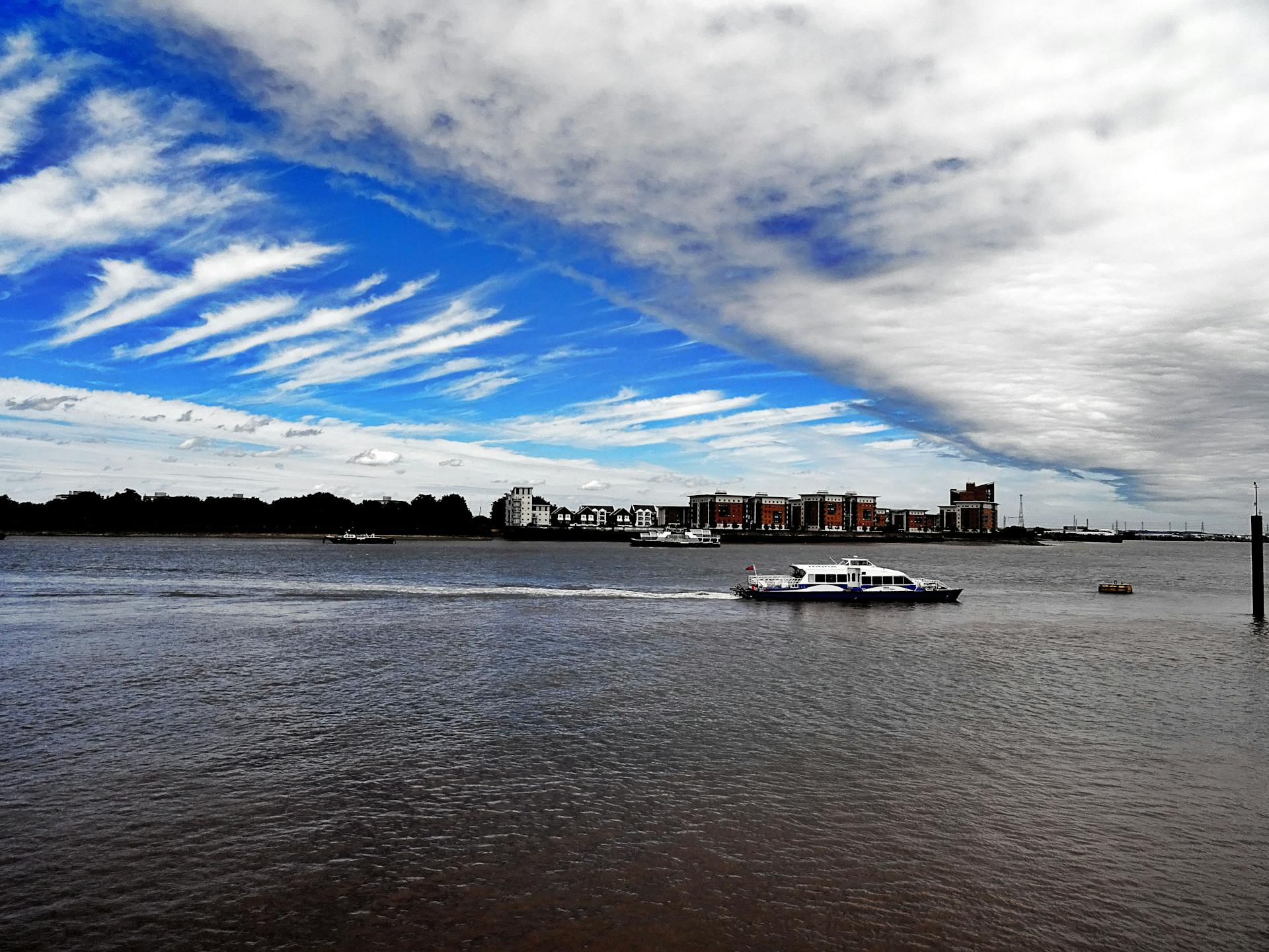 A vibrant urban scene with a ferry traversing a wide river beneath striking cloud formations.
