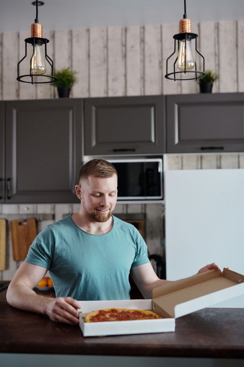 Free A cheerful man in a modern kitchen opens a pizza box, revealing a delicious meal. Stock Photo