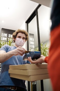 A deliveryman wearing a mask delivers pizza and completes a contactless payment indoors.