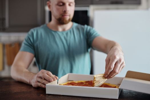 A man in a teal shirt happily taking a slice from a pizza box on a wooden table.