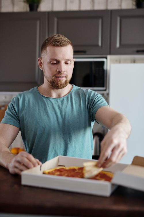 Free Adult man at home kitchen taking a slice from a pizza box. Casual indoor scene. Stock Photo