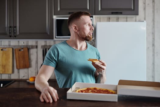 A man in a casual t-shirt enjoys pizza in a stylish kitchen environment.