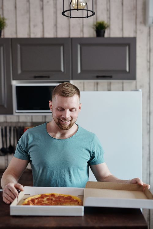 Free Cheerful man opening a pizza box in a modern kitchen, ready to enjoy his meal. Stock Photo