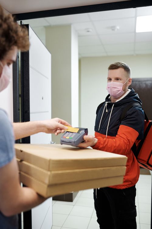 Free Deliveryman in mask handing pizza and credit card machine to a customer indoors. Stock Photo
