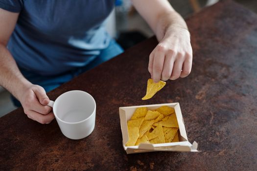 Close-up of a man holding nachos and a ceramic cup, emphasizing casual snack time.