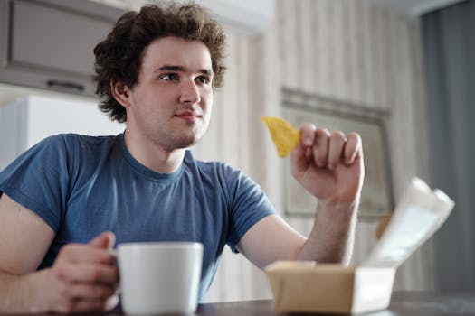 Casual young man in a blue t-shirt enjoying nachos with a mug in a modern kitchen setting.