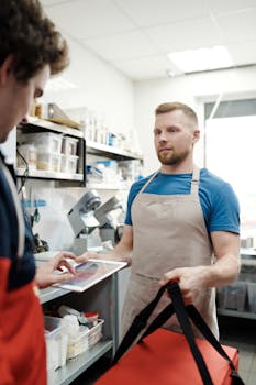Man in apron holding thermal food bag preparing a delivery order in a restaurant kitchen.