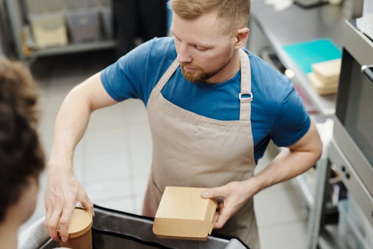 Man In Apron Putting Box And Cup In Bag