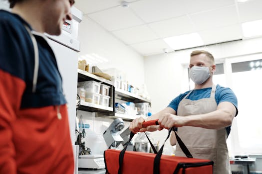 A male food delivery worker in an apron and face mask handling a thermal bag.