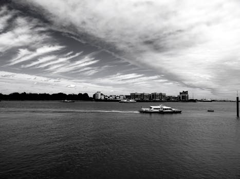 Black and white photo showcasing a serene seascape with a modern city in the background under dramatic clouds.