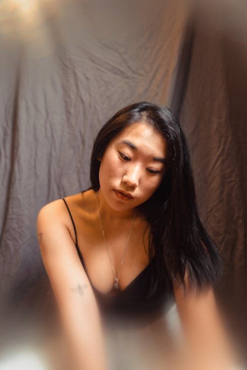 Emotionless Asian female in tank top sitting on floor in studio on black background and looking down
