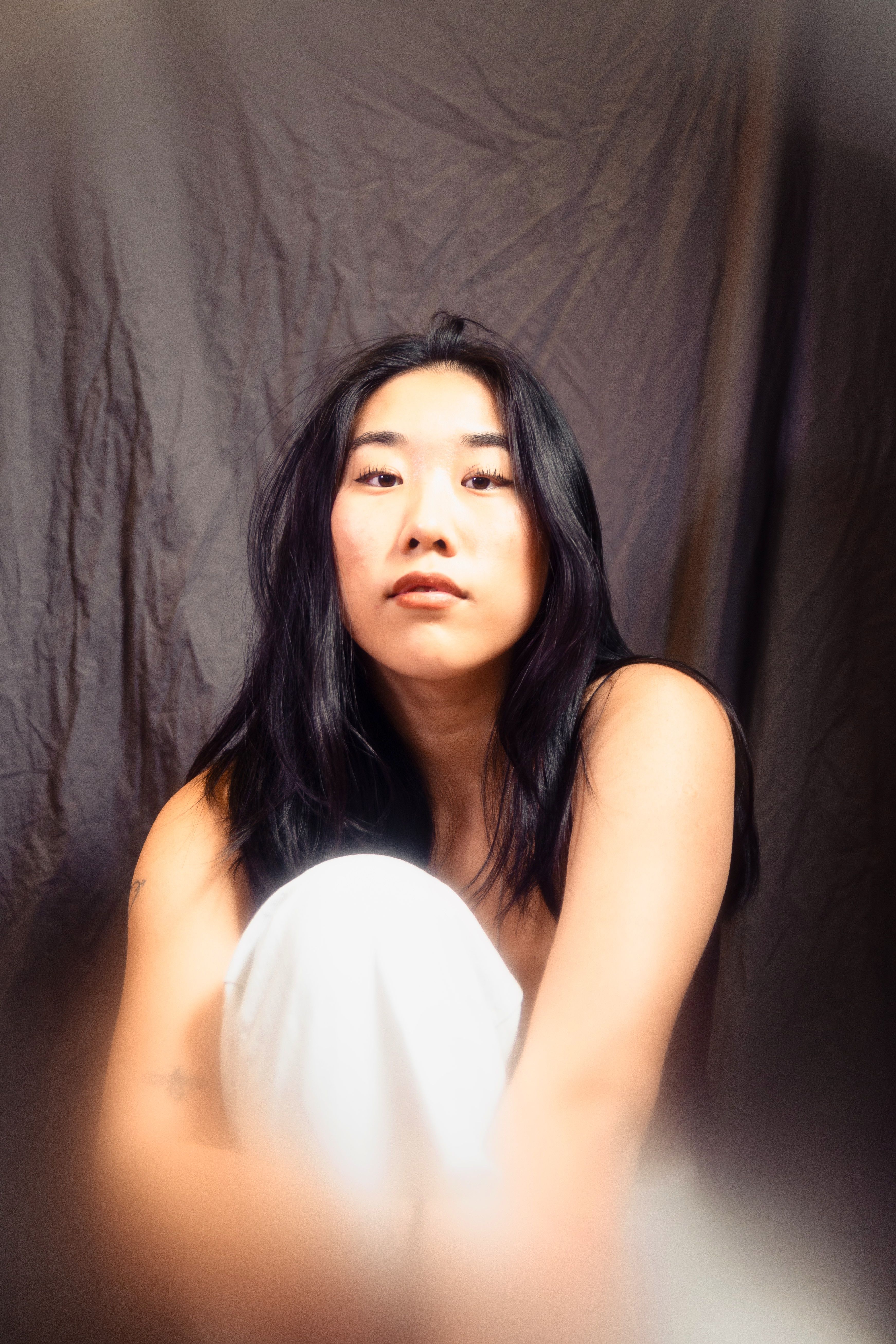 Peaceful Asian female model sitting on floor in studio on black background and embracing knee while looking at camera