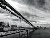 这是一张黑白照片，画面中一座工业桥横跨河流，背景是城市天际线