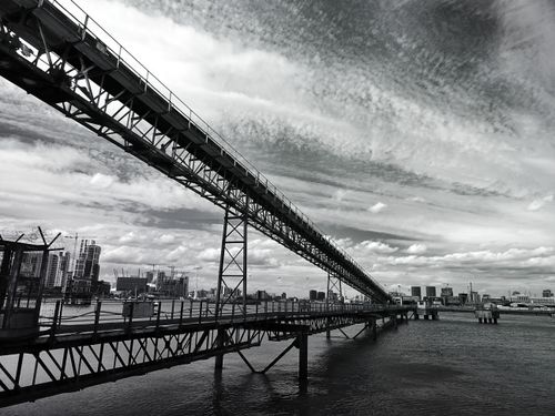 Free Black and white photo of an industrial bridge spanning across the river with an urban skyline in the background. Stock Photo