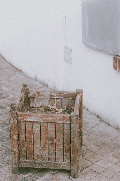 Garbage in small wooden square shabby dirty basket on pavement next to old white wall on street