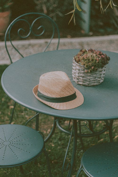 Beige straw hat next to flower basket on dark green round table surrounded by stools with back rest decorated with ornaments in outside cafe