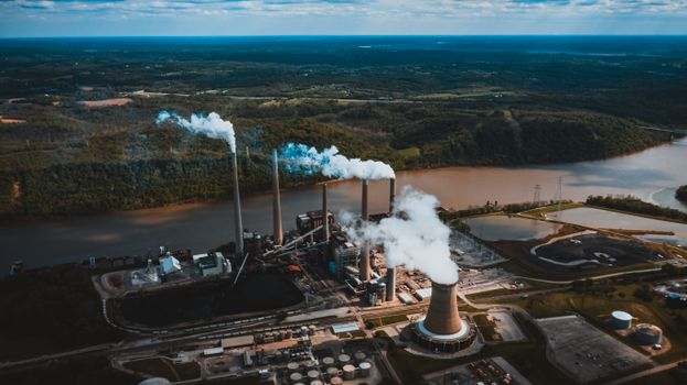 Aerial view of industrial landscape of city with smoke emissions from chimneys located near river and forest