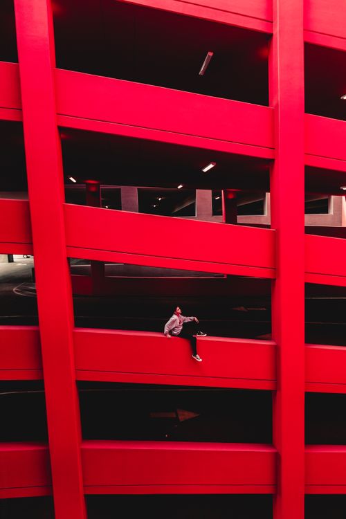 Free Individual sitting in a red multi-story parking structure, showcasing urban architecture. Stock Photo