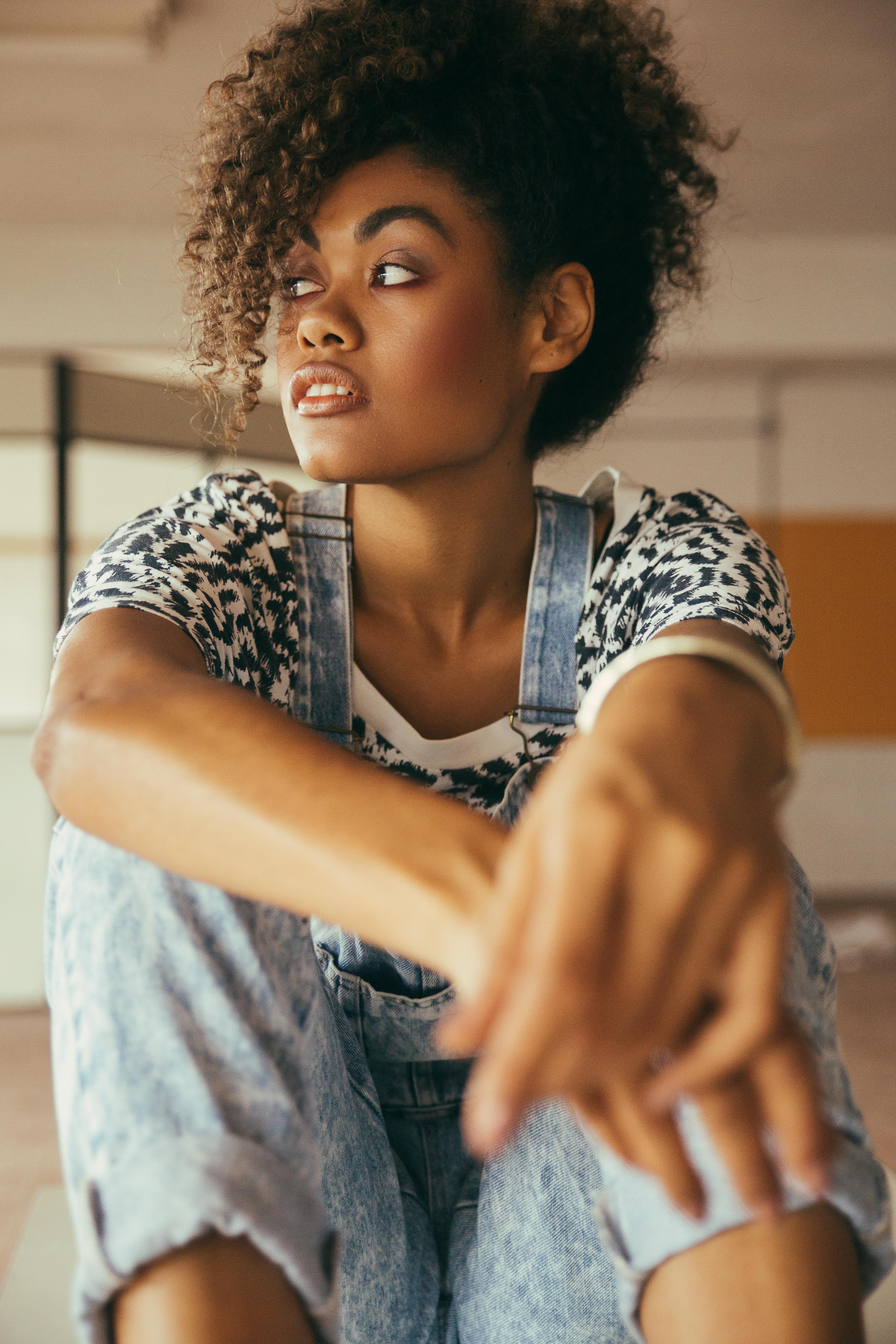 Crop pondering African American lady with makeup in stylish denim overalls looking away in building