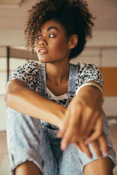 Crop pondering African American lady with makeup in stylish denim overalls looking away in building