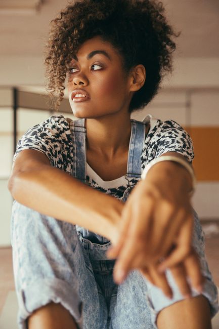 Crop pondering African American lady with makeup in stylish denim overalls looking away in building