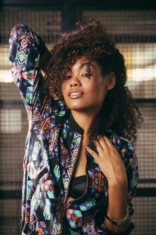 Young feminine black female in stylish wear with curly hair standing with raised arm while looking at camera