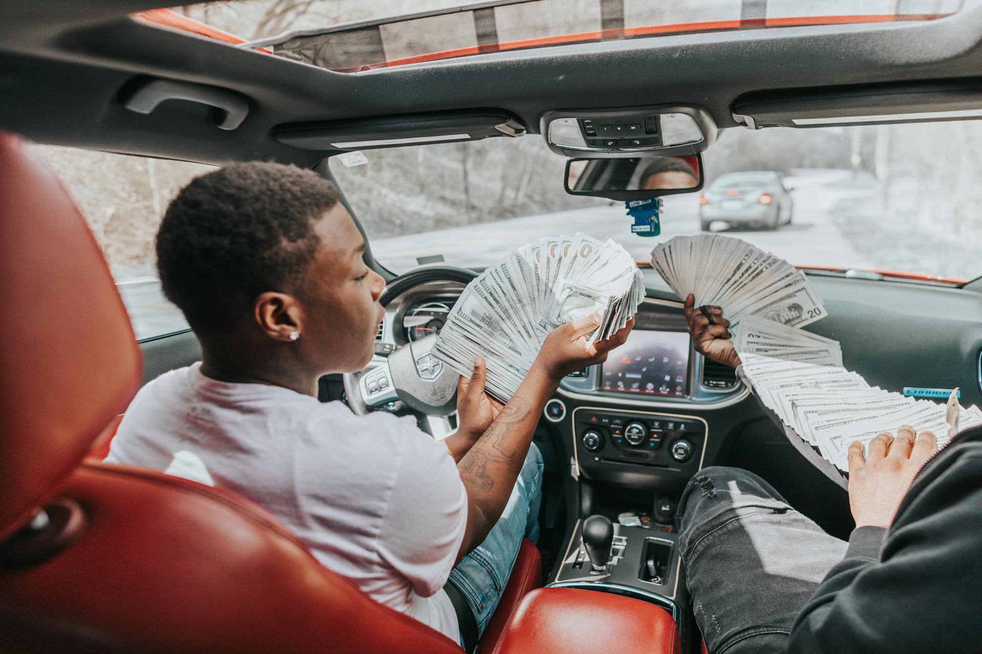 Two men displaying large amounts of cash inside a luxury car, highlighting wealth and lifestyle.