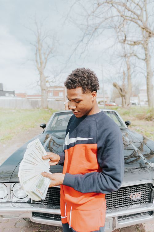 Free A young man stands outside with a classic car, holding a fan of cash. Stock Photo
