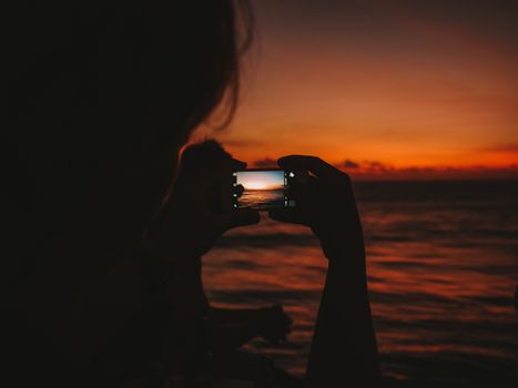 Silhouette shot of a person capturing a vibrant Bali sunset over the ocean with a smartphone.