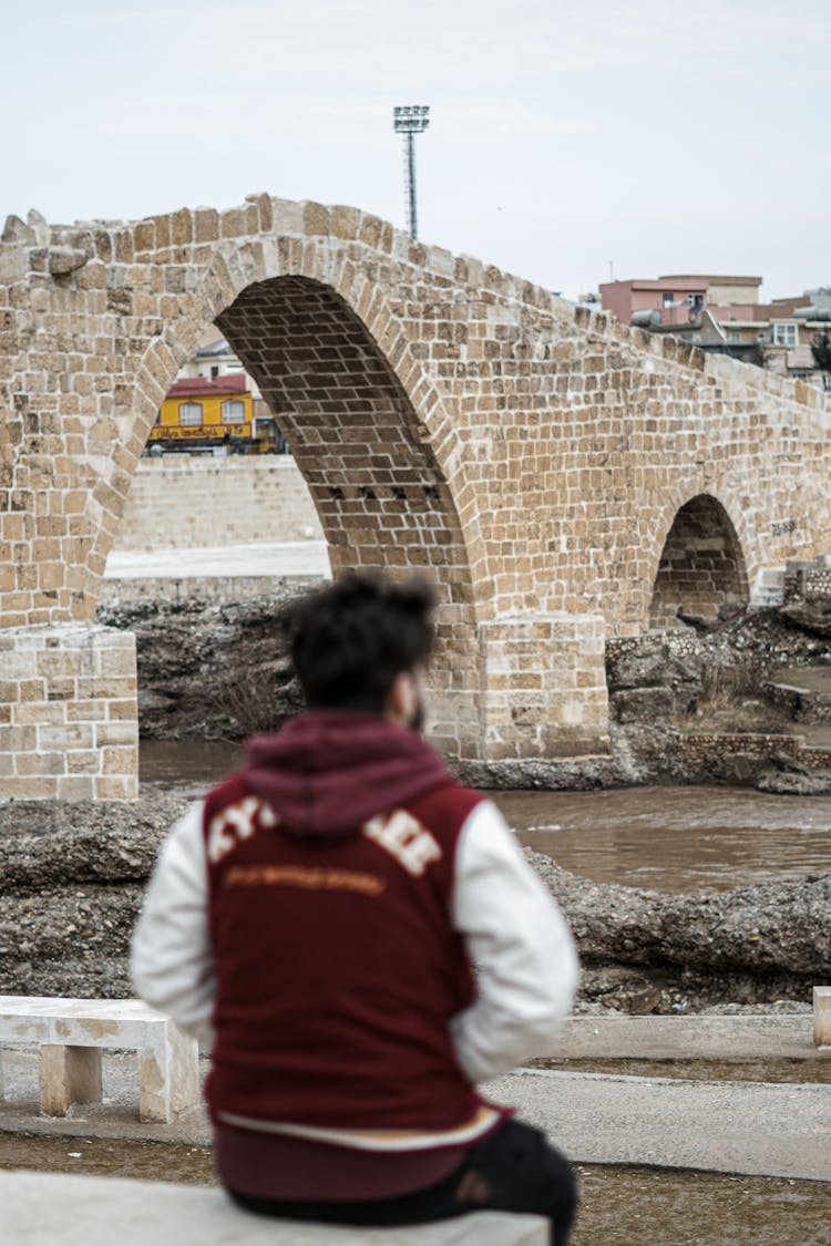 Man In Red And White Hoodie Sitting Near A Stone Arch Bridge