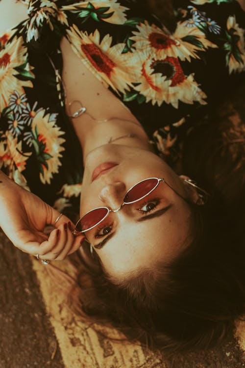 Free From above of trendy female in bright wear with makeup and accessories touching sunglasses while looking at camera and lying on road Stock Photo