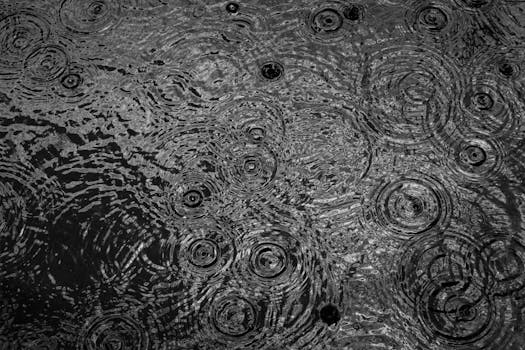 Black and white photo showcasing ripples on a puddle surface during rainfall, creating a textured pattern.