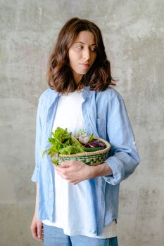 Caucasian woman holds a basket of fresh vegetables, looking contemplative. Indoors, neutral background.
