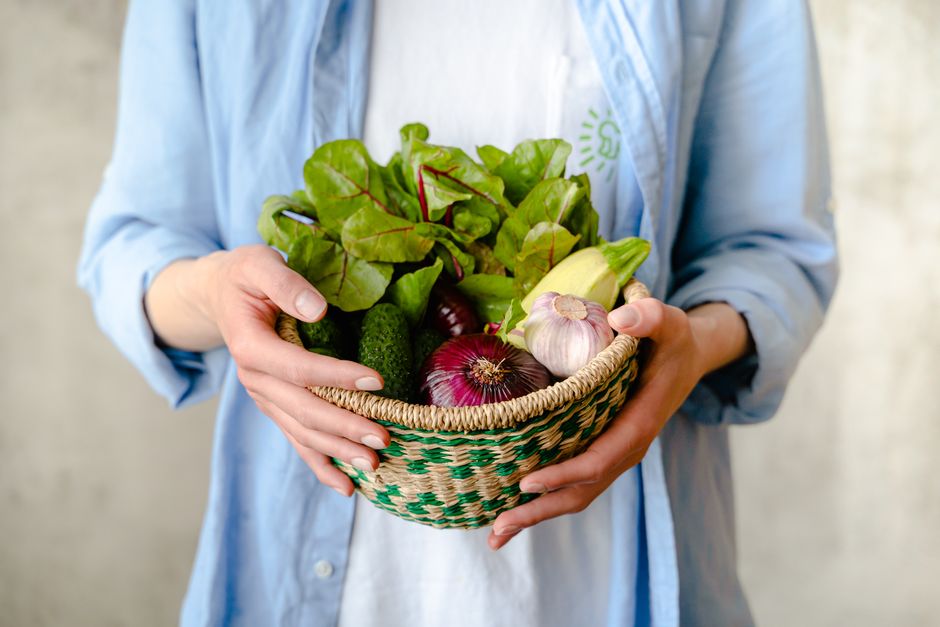 Hands holding a basket filled with fresh vegetables and herbs.