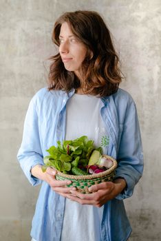 Caucasian woman in casual attire holding a basket of fresh vegetables indoors.