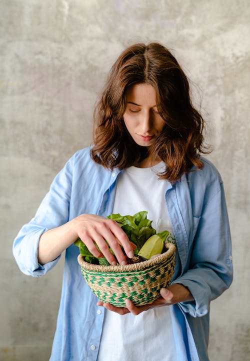 Woman in Blue Long Sleeve Shirt Holding Brown Woven Basket With Green Vegetable