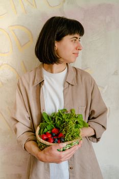 A woman holding a woven basket filled with fresh berries and greens, looking thoughtful indoors.