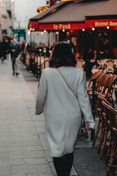 黄昏时分，一名穿着外套的妇女漫步在城市街道的咖啡馆旁，温暖的灯光闪烁