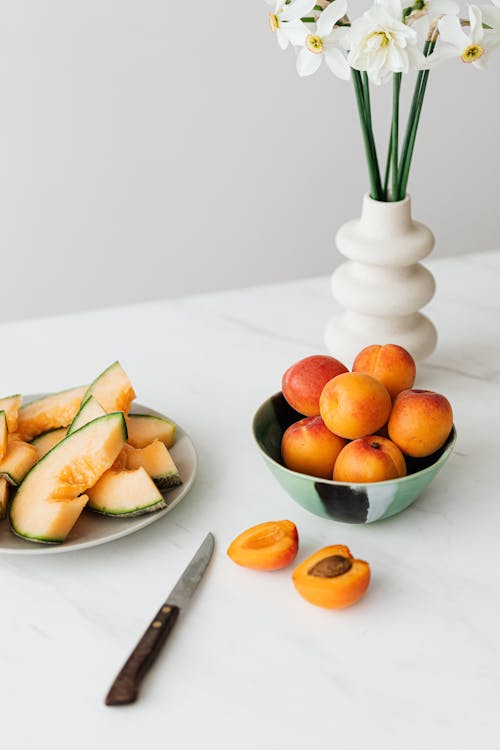 From above bowl of fresh ripe apricots and sliced melons composed with white vase of beautiful narcissus on table