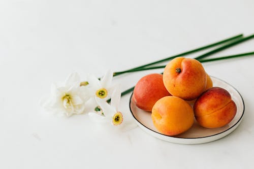 Free From above of appetizing ripe apricots in ceramic plate composed with fresh delicate white narcissus flowers Stock Photo