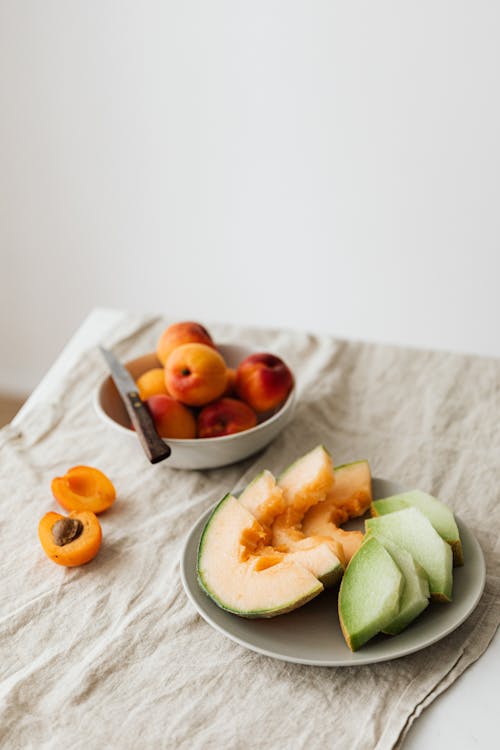Free Still life of delicious healthy assorted melons on plate and bowl with fresh ripe apricots placed on linen tablecloth Stock Photo