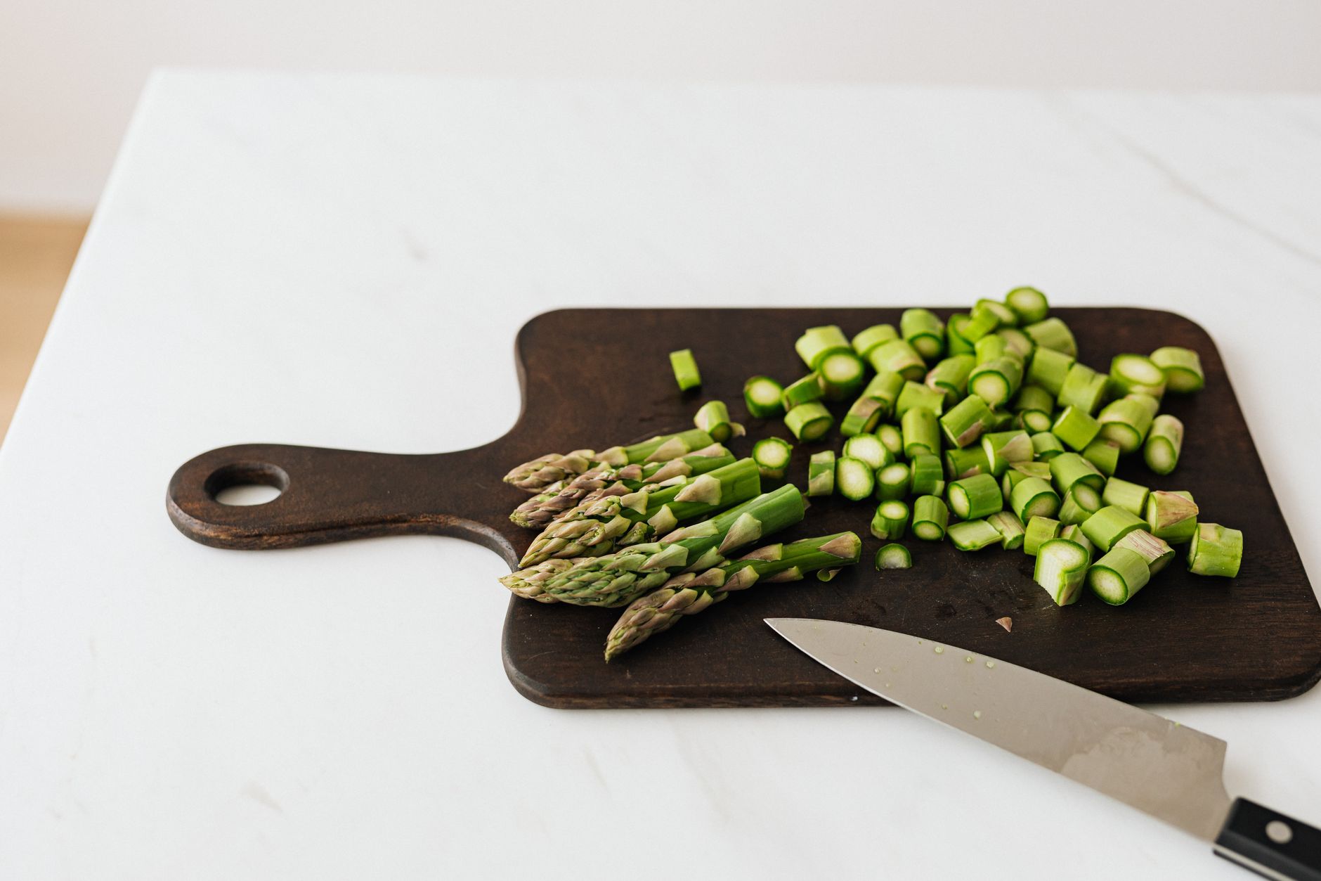 From above of chopped green asparagus near knife on wooden cutting board placed on white table