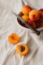 From above of ceramic bowl with ripe colorful fruits and knife placed on white rumpled  tablecloth near cut peach in half