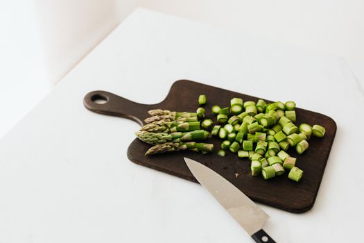 From above of chopped fresh asparagus near knife placed on dark cutting board composed on white table in kitchen
