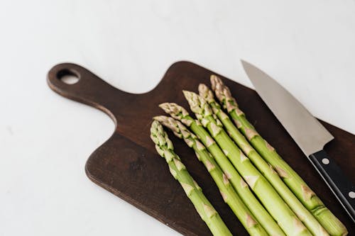 From above of green asparagus placed near kitchen knife on wooden cutting board on white table