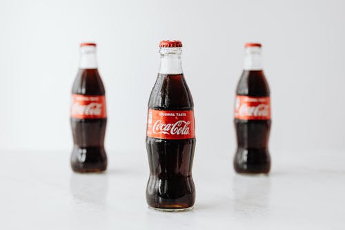 Modern glass bottles of cold soda with red label placed on white reflective surface