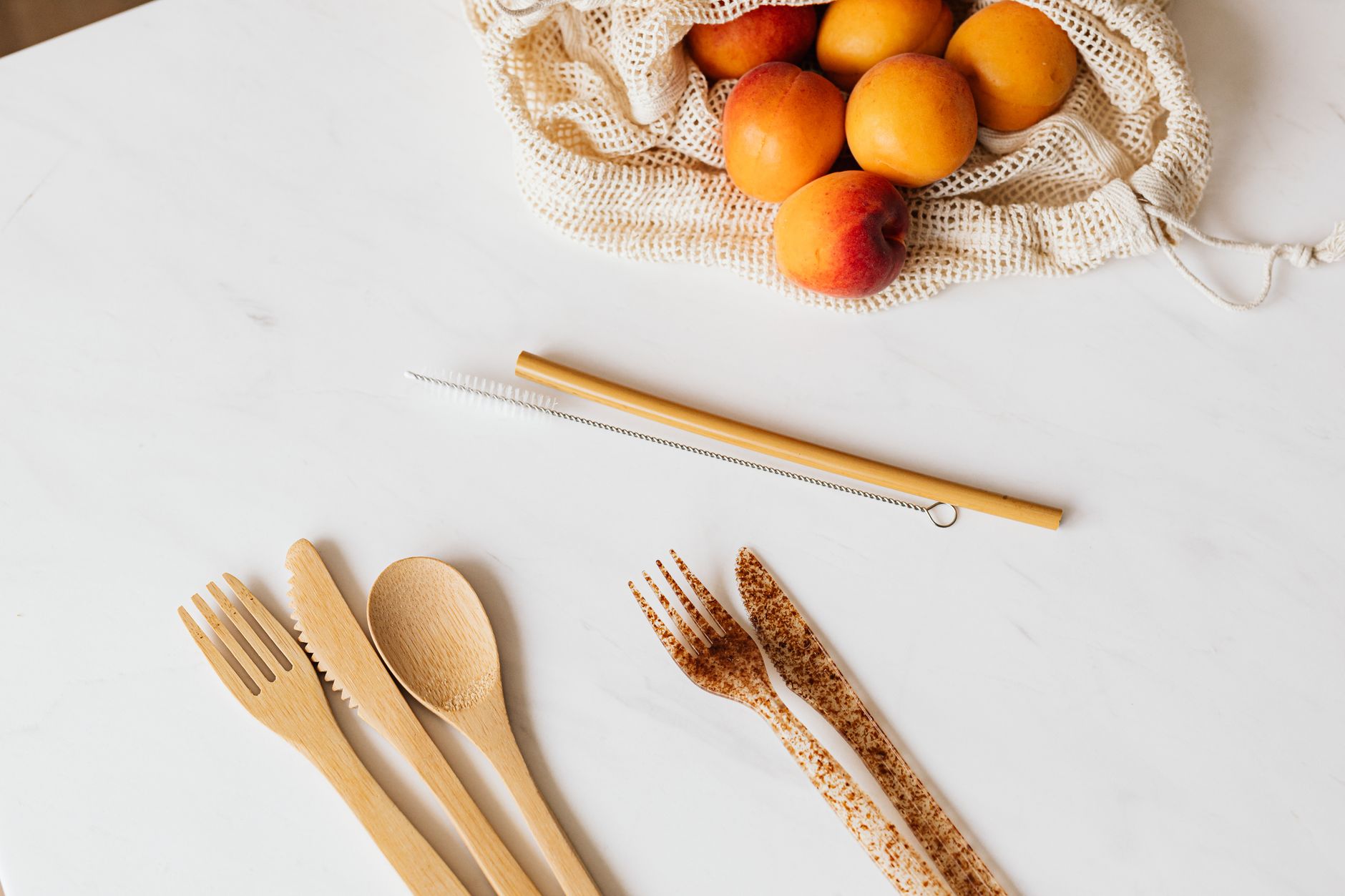 From above composition of cutlery made from different materials placed on white table with colorful peaches in fabric bag