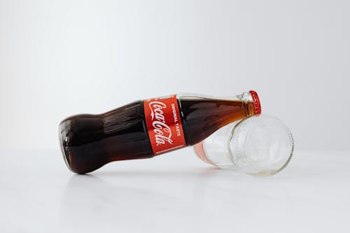 Closed bottle of soft drink leaned on empty bottle lying on white reflective surface