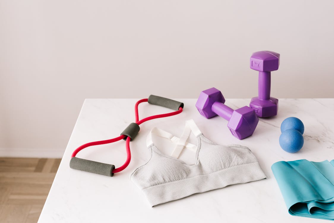 Free From above of white sports bra surrounded by purple dumbbells and blue tape expander and red tubular expander and blue massage double ball placed on marble surface of table Stock Photo